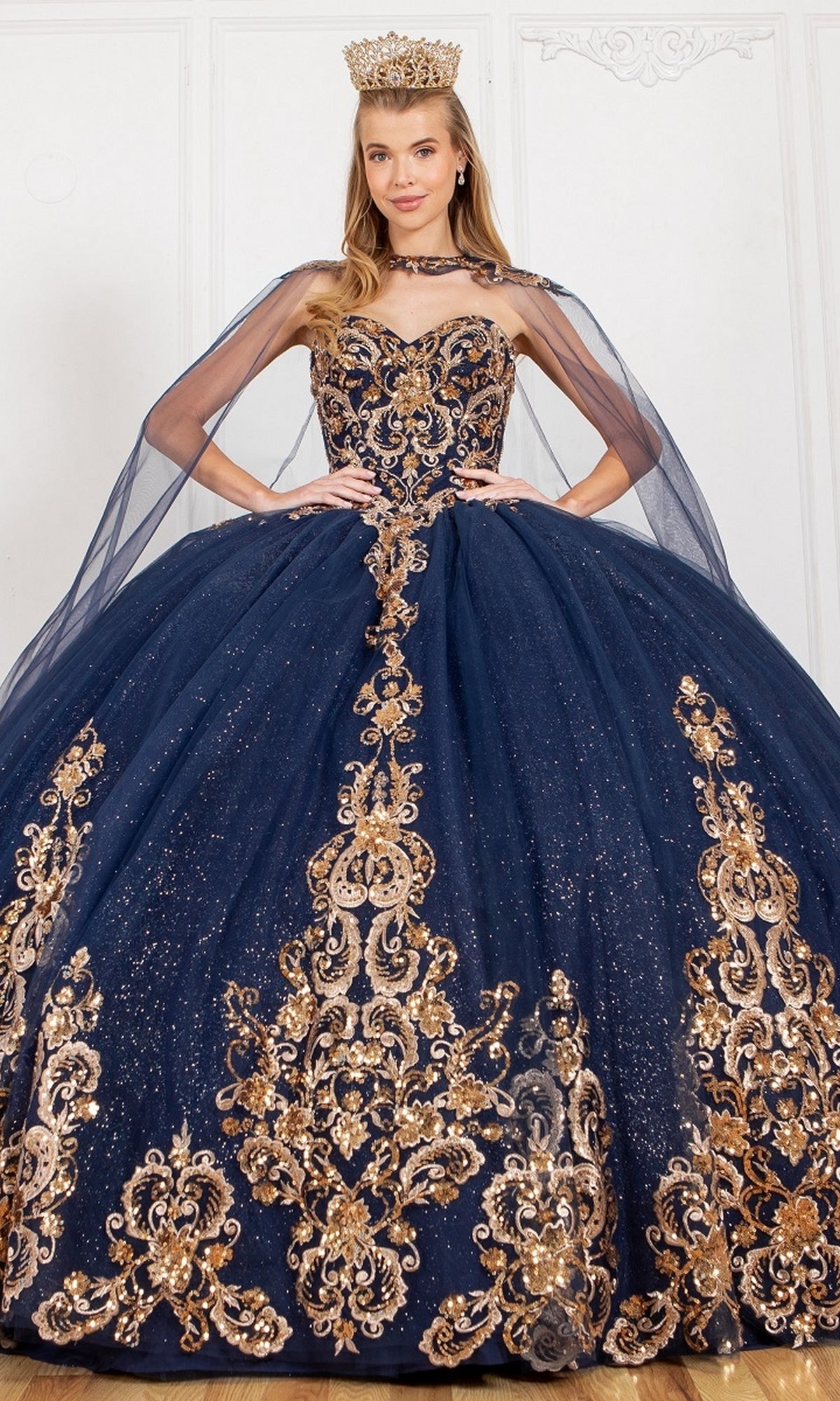 A woman models the Quinceanera Ball Gown 8063J—a navy blue ball gown with gold embroidery, glitter tulle, sheer cape sleeves, and a gold crown—standing indoors against a white background.